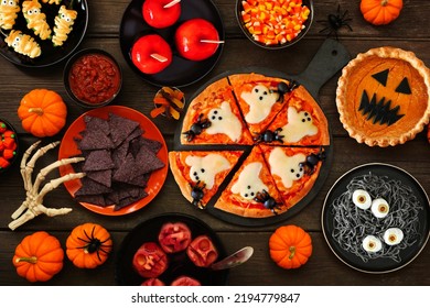 Fun Halloween Dinner Party Table Scene Over A Dark Wood Background. Overhead View. Pizza, Jack O Lantern Pumpkin Pie, Candy Apples, Eyeball Spaghetti, Snacks And Spooky Punch.