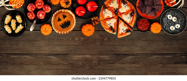 Fun Halloween Dinner Party Top Border Over A Dark Wood Banner Background. Overhead View. Pizza, Jack O Lantern Pumpkin Pie, Candy Apples, Eyeball Spaghetti, Snacks And Spooky Punch.