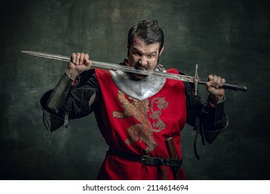 Fun, joy. Comic portrait of funny medieval warrior or knight with dirty wounded face holding sword isolated over dark background. Comparison of eras, history, renaissance style. Art, meme