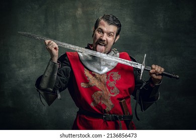 Fun, Joy. Comic Portrait Of Funny Medieval Warrior Or Knight With Dirty Wounded Face Holding Sword Isolated Over Dark Background. Comparison Of Eras, History, Renaissance Style. Art, Meme