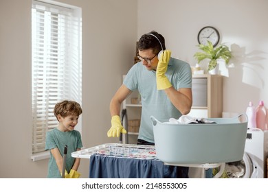 Fun In The Laundry Room. Dad And Son Doing Household Chores, Fooling Around While Cleaning. Man Pretends To Play DJ On Dryer, Child Holds Mop As Guitar, Musical Concert.