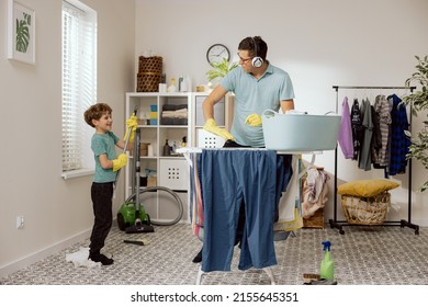 Fun In The Laundry Room. Dad And Son Doing Household Chores, Fooling Around While Cleaning. Man Pretends To Play DJ On Dryer, Child Holds Mop As Guitar, Musical Concert.