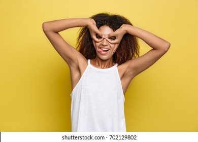 Fun And People Concept - Headshot Portrait Of Happy Alfo African American Woman With Freckles Smiling And Making Finger Glasses. Pastel Yellow Studio Background. Copy Space.