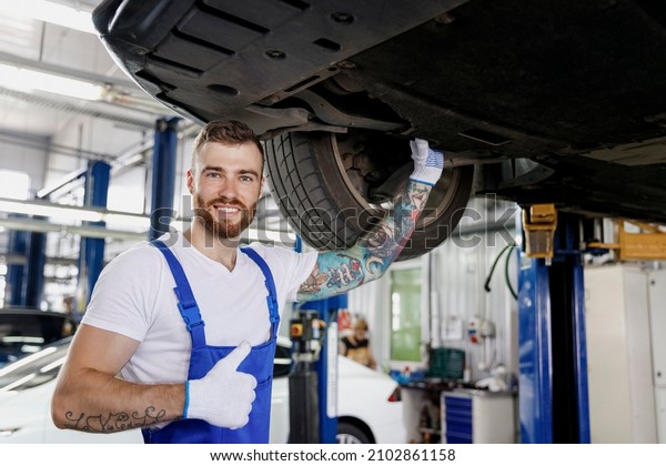 Fun repairman young male professional technician\
mechanic man in denim blue overalls t-shirt show thumb up stand\
near car lift check technical condition work in vehicle repair shop\
workshop indoors