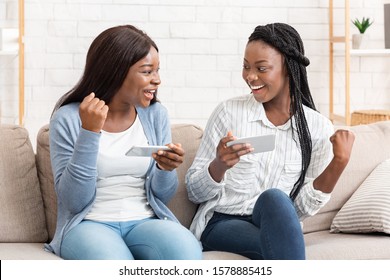 Fun With Sister. Two Afro Girls Playing Video Games On Smartphones And Celebrating Victory With Clenched Fists, Sitting On Couch At Home.