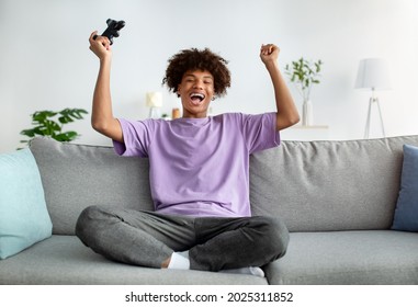 Fun Stay Home Activities. Overjoyed Black Teenager With Joystick Winning Online Video Game, Gesturing YES And Screaming In Excitement On Couch Indoors, Full Length Portrait