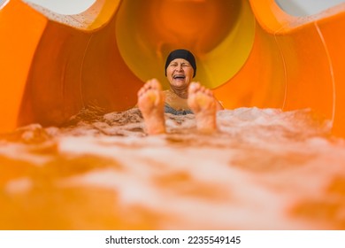 Fun swimming pool or aqua park activities. Laughing amused elderly caucasian female pensioner in black hair cap sliding on an orange water slide. Blurred feet in the foreground. High quality photo