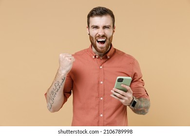 Fun tatooed young brunet man 20s short haircut with earrings wear apricot shirt hold in hand use mobile cell phone doing winner gesture celebrating isolated on pastel orange background studio portrait