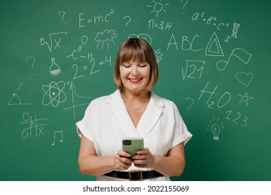 Fun Teacher Mature Elderly Senior Lady Woman 55 Wear White Shirt Hold In Hand Use Mobile Cell Phone Isolated On Green Wall Chalk Blackboard Background Studio. Education In School September 1 Concept