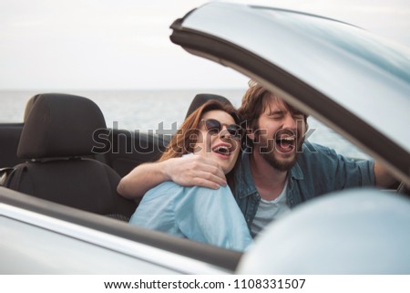 Fun together. Joyful happy couple in love is sitting in luxury cabriolet and hugging. They are driving car along seaboard and laughing. Male and female are relishing summer vacation abroad