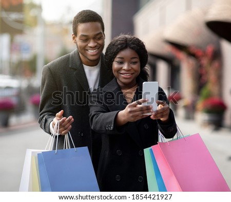 Fun together and selfies during Black Friday and season sale. Smiling millennial african american guy and woman with colored bags make selfie outdoors, near shopping mall for social media