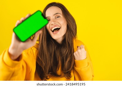Fun woman hold in hand, point on phone green screen doing winner gesture clenching fists isolated on yellow background. Girl using smartphone apps winning online celebrating discount gift voucher. - Powered by Shutterstock