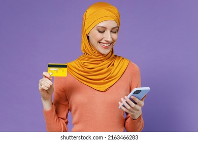 Fun Young Arabian Asian Muslim Woman In Abaya Hijab Yellow Clothes Using Mobile Cell Phone Hold Credit Bank Card Doing Online Shopping Isolated On Plain Pastel Light Violet Background Studio Portrait