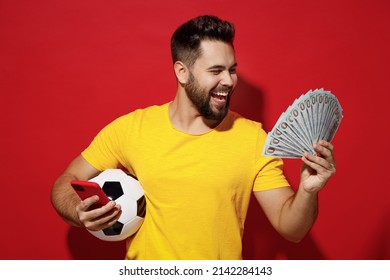 Fun young bearded man football fan in yellow t-shirt cheer up support favorite team hold soccer ball look at fan of cash money in dollar banknotes isolated on plain dark red background studio portrait - Shutterstock ID 2142284143