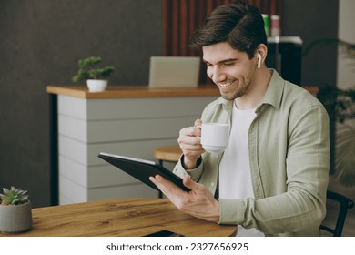 Fun young man wear casual clothes sits alone at table in coffee shop cafe indoors work or study use digital tablet pc computer listen music in earphones drink tea. Freelance mobile business concept
