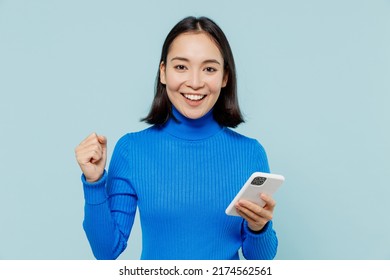 Fun Young Woman Of Asian Ethnicity 20s Years Old Wears Blue Shirt Hold In Hand Use Mobile Cell Phone Doing Winner Gesture Clenching Fists Isolated On Plain Pastel Light Blue Background Studio Portrait
