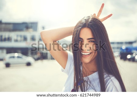 Fun young woman making a horns gesture behind her head with her fingers as she smiles at the camera outdoors in town