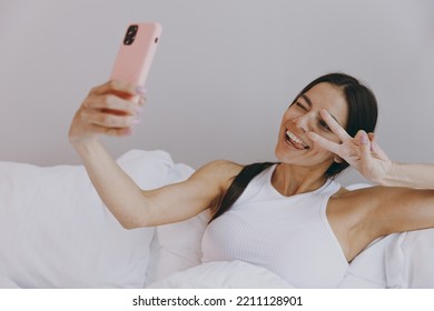 Fun Young Woman In White Tank Top Sit In Bed Get Video Call Using Mobile Cell Phone Doing Selfie Talk Showing Victory Sign In Bedroom Lounge Home In Own Room House Wake Up Be Lost In Reverie Good Day