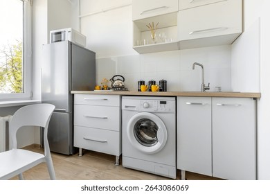 Functional kitchen interior with washing machine, sink, kettle, mugs, modern appliances and wooden countertop in a bright home.