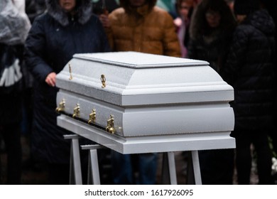 Funeral Ceremony. Grieving People Look At A White Wooden Coffin In The Rain In A Cemetery.