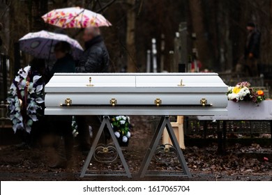 Funeral Ceremony. Grieving People Are Standing In The Rain In A Cemetery With Umbrellas In Their Hands Near A White Coffin.