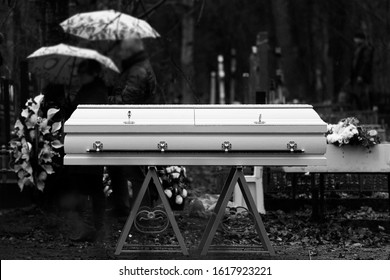 Funeral Ceremony. Grieving People Are Standing In The Rain In A Cemetery With Umbrellas In Their Hands Near A White Coffin.  Black And White Photo