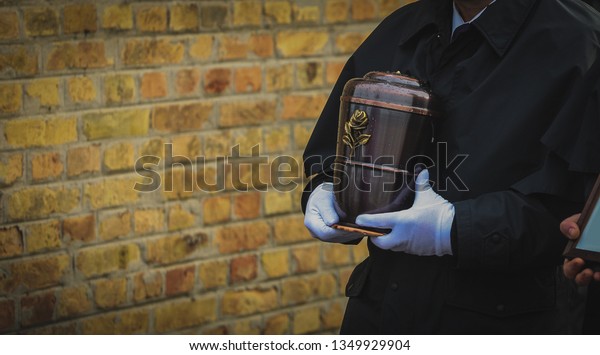 Funeral director or undertaker\
carrying a smooth metal urn in his hands, filled with ash towards\
the grave. Brick wall in the background. Funeral\
procession