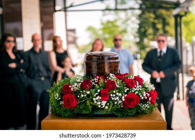 Funerary urn with ashes of dead and flowers at funeral. Burial urn decorated with flowers and people mourning in background at memorial service, sad and grieving last farewell to deceased person. - Shutterstock ID 1973428739