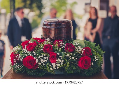 Funerary Urn With Ashes Of Dead And Flowers At Funeral. Burial Urn Decorated With Flowers And People Mourning In Background At Memorial Service, Sad And Grieving Last Farewell To Deceased Person.