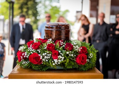 Funerary Urn With Ashes Of Dead And Flowers At Funeral. Burial Urn Decorated With Flowers And People Mourning In Background At Memorial Service, Sad And Grieving Last Farewell To Deceased Person.