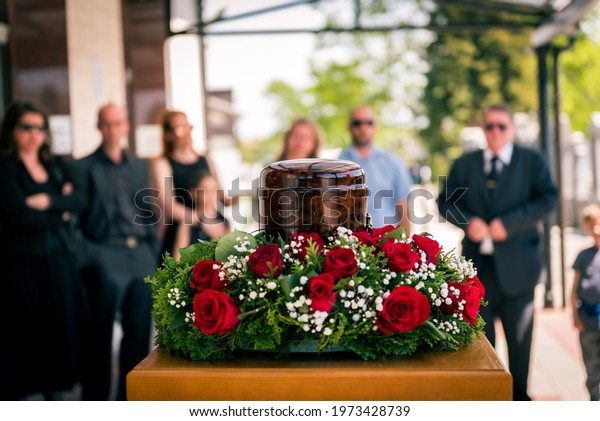 Funerary urn with ashes of dead and flowers at\
funeral. Burial urn decorated with flowers and people mourning in\
background at memorial service, sad and grieving last farewell to\
deceased person.