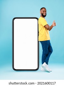 Funky black guy leaning on giant cellphone, showing thumb up gesture, recommending new mobile app or cool website, mockup for your ad design. Blue studio background