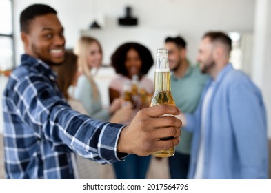 Funky Black Guy Showing Bottle Of Beer, Celebrating With Group Of His Diverse Friends, Saying CHEERS At Home, Selective Focus. Young Students Drinking Alcohol, Having Social Gathering Or Party