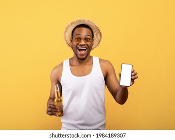 Funky black guy in summer clothes holding cellphone with blank screen and bottle of beer on yellow background, mockup for mobile app or website. Young man with empty smartphone drinking alcohol
