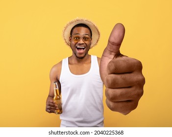 Funky Black Guy In Summer Clothes Holding Bottle Of Beer And Showing Big Thumb Up On Yellow Studio Background, Mockup For Mobile App Or Website. Young Afro-american Man Drinking Alcohol, Having Fun