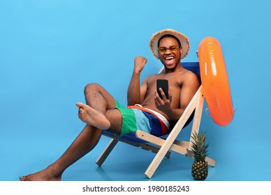 Funky black guy in swinwear holding smartphone, feeling excited, sitting in lounge chair, gesturing YES on blue studio background. African American man learning about huge sale or discount, copy space
