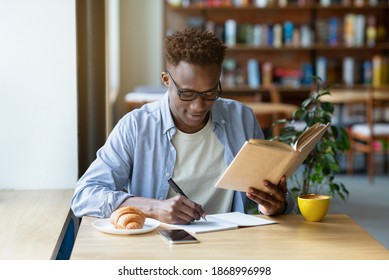 Funky Black Guy With Textbook Taking Notes In Copybook At Cozy Cafe. Positive African American Man Studying At Coffee Shop, Reading Book, Writing Down Information, Getting Ready For Exam