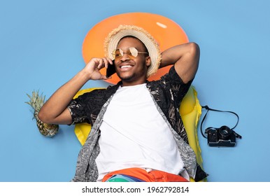 Funky black guy in trendy summer clothes lying on inflatable lilo, speaking on smartphone over blue studio background, top view. African American man having phone conversation on beach vacation