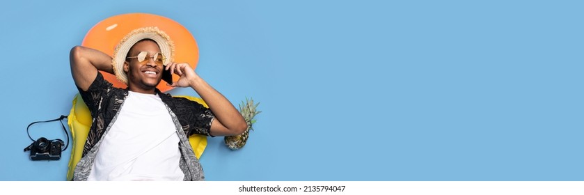 Funky black guy in trendy summer clothes lying on inflatable lilo, speaking on smartphone over blue studio background, top view. Man having phone conversation on beach vacation, banner, copy space - Powered by Shutterstock