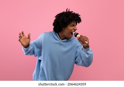 Funky Black Teenager Singing Song Into Microphone, Performing Karaoke On Pink Studio Background. Cool African American Teen Guy With Mic Pretending To Be Rock Star