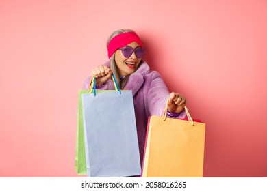 Funky And Cool Asian Senior Woman In Fashionable Clothes Dancing While Going Shopping On Sales, Holding Shop Paper Bags And Having Fun, Pink Background