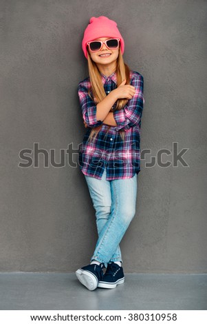 Funky girl. Full length of cheerful little girl in sunglasses keeping arms crossed and looking at camera with smile while standing against grey background