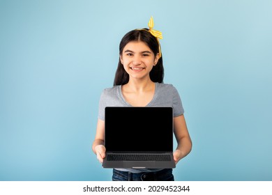 Funky Indian Teen Girl Holding Laptop Pc With Blank Screen For Your Ad Or Website On Blue Studio Background. Cool Asian Adolescent Offering Mockup For Design Or Advertisement