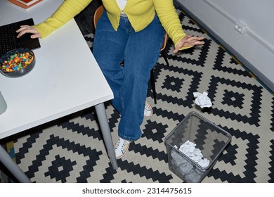 Funky top view shot of young woman having fun at workplace and throwing paper into basket, copy space