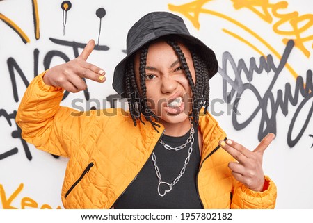 Funky urban teenage girl with dreadlocks makes yo gesture poses against graffiti artwork dressed in fashionable clothes or street fashion outfit feels cool. Teenagers lifestyle and free time concept