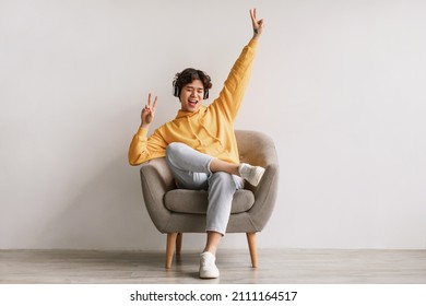 Funky young Asian man listening to music in wireless headphones, dancing, sitting in armchair against white studio wall, full length. Handsome millennial male enjoying favorite playlist