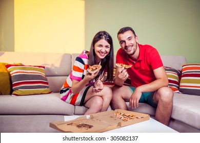 Funky Young Couple Eating Pizza On A Couch In Front Of A Green Wall