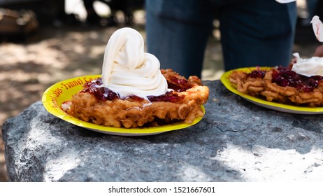 Funnel Cake With Whipped Cream And Strawberry Compote On Top Sitting On A Yellow Paper Plate.