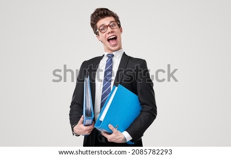 Funny accountant in nerd glasses and formal suit standing with folders of reports and opened mouth in studio against gray background