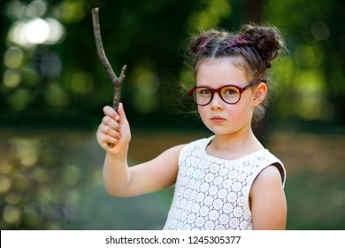 Funny Adorable Little Kid Girl With Glasses And Wooden Magic Wand Playing Harry Potter In Park On Sunny Day. Fan Of Magic Story.